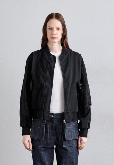 Fekete bomberdzseki, szűkített derékkal, oldalsó zsebekkel és bordázott mandzsettákkal. Sima textúrájú, elöl cipzárral záródik. Fehér felső látható alatta.