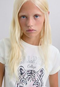 Tricou alb cu un grafic de tigru, ornamentat cu strasuri. Mâneci scurte, guler rotund, croială lejeră, purtat de un model cu păr lung și deschis la culoare.
