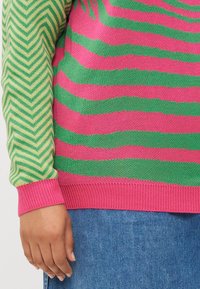 Knit sweater featuring bold pink and green horizontal stripes, with a contrasting green and yellow chevron pattern on one sleeve, ribbed cuffs.