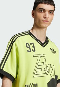 Šviesiai geltona sportinė marškinėliai su juodais akcentais, V formos iškirpimu ir drąsiais grafikos elementais. Priekyje yra spausdintas numeris "93" ir žodžiai "TEAM".