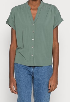 Frau trägt grüne Kurzarmbluse mit hochgekrempelten Ärmeln, kombiniert mit hoch taillierter blauer Jeans, steht vor hellem Hintergrund.