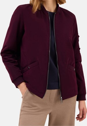 Mujer con una chaqueta bomber de color púrpura oscuro con cremallera y bolsillos, y pantalones beige, de pie con una mano en el bolsillo.