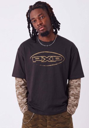 Jeune homme avec des dreadlocks portant un t-shirt noir avec le logo PXP par-dessus des manches et un pantalon camouflage, debout les mains dans les poches sur un fond uni.