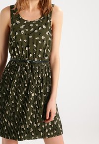 Vestido verde sin mangas con un patrón de estampado de leopardo, con una falda plisada y un cinturón negro a la cintura. Textura de tela ligera.