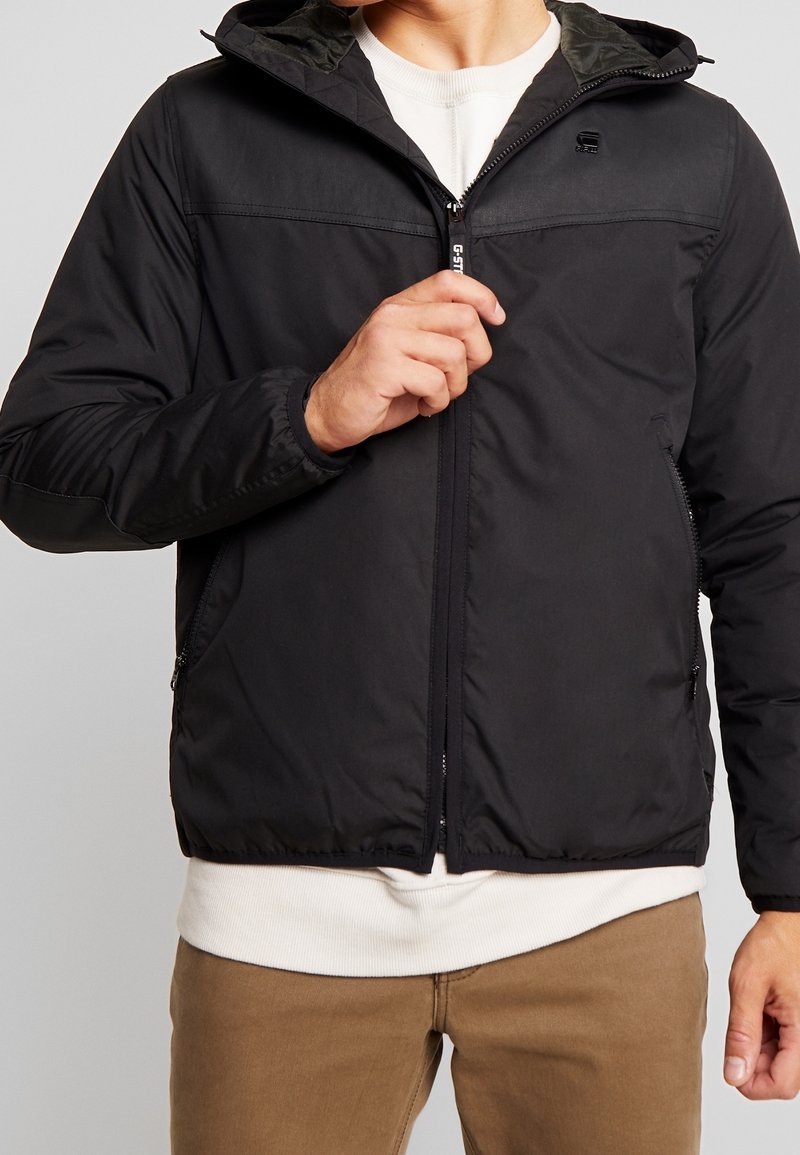 Veste noire zippée avec capuche, présentant une surface extérieure lisse, des poches latérales et un logo subtil sur la poitrine. Doublée d'un matériau contrastant.