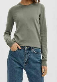 Pull en maille verte avec un col arrondi festonné et des poignets côtelés, associé à un jean en denim bleu taille haute.