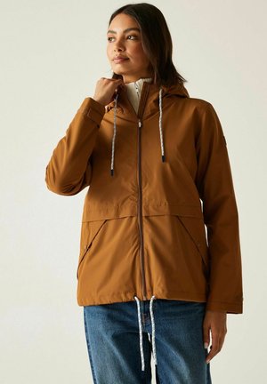 Woman wearing brown hooded jacket with white drawstrings and blue jeans, looking slightly to the side with hand near collar.