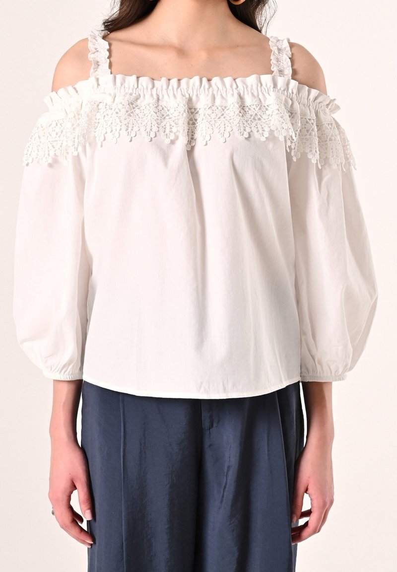 Blouse blanche épaules dénudées avec bordure en dentelle et bretelles à volants, associée à un pantalon ample bleu foncé, présentée des épaules aux hanches.