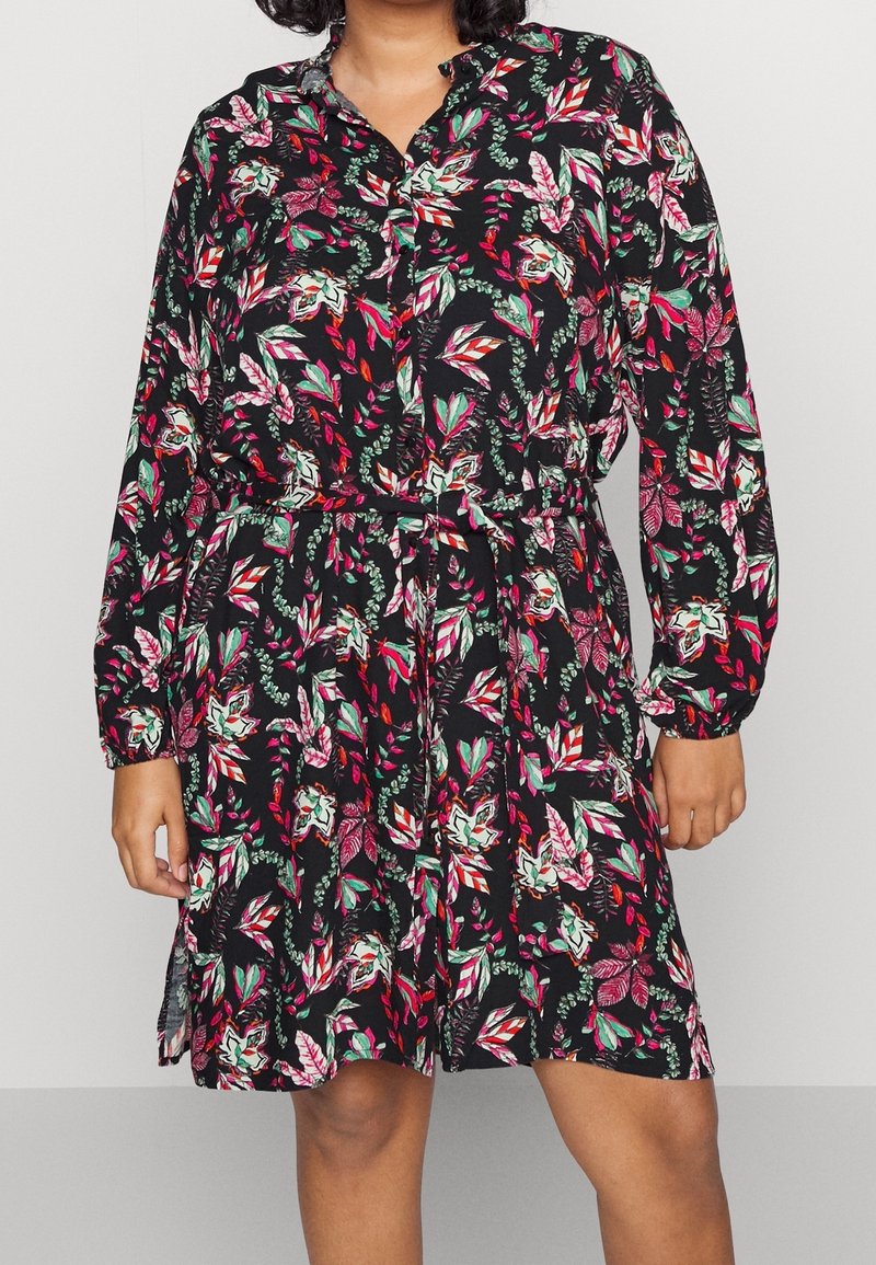 Femme portant une robe noire à motifs floraux rouges, verts et blancs, manches longues, boutons et ceinture nouée à la taille.