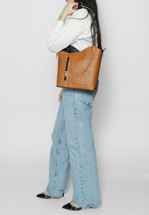 Bruine leren emmerzak met een geperforeerd stippenpatroon, enkele ritssluiting en zwarte schouderband, gecombineerd met lichtblauwe flare jeans.
