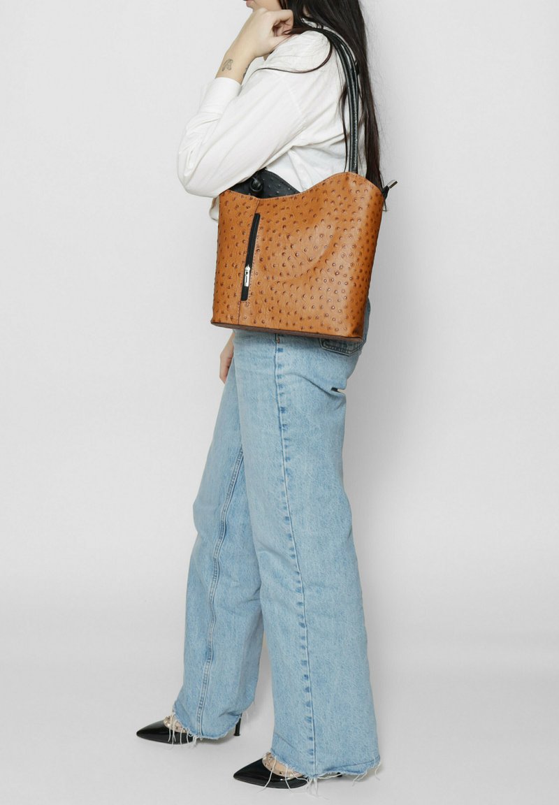 Bruine leren emmerzak met een geperforeerd stippenpatroon, enkele ritssluiting en zwarte schouderband, gecombineerd met lichtblauwe flare jeans.