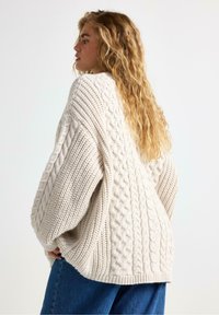 Kabelstrikket sweater i off-white, med en struktureret overflade, voluminøse ærmer og en afslappet pasform. Sammen med mørkeblå jeans.