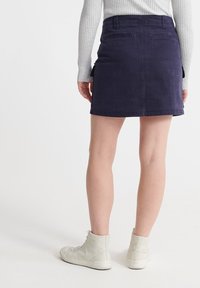 Person iført en ribbet langærmet bluse, en marineblå miniskørt med lommer og hvide høje sneakers, stående foran en ensfarvet baggrund.