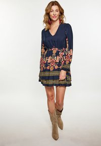 Rochie bleumarin cu broderie florală în roșu, galben și alb; mâneci lungi; talie strânsă; tiv cu volane în straturi; asociată cu cizme cowboy bej.