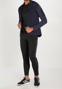 Granatowa kurtka sportowa z wzorzystymi rękawami, kieszeń zapinana na zamek, czarna koszulka, czarne spodnie ze niebieskimi paskami i czarne tenisówki z szarymi akcentami.