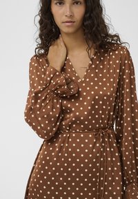 Rjavo obarvana obleka s pikami, z V-izrezom, dolgimi nabranimi rokavi in zavezovanjem v pasu. Tkanina je videti gladka in lahka s prijetnim sijajem.