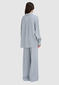 Jasnoszara, luźna koszula z długim rękawem i plisą z tyłu oraz mankietami zapinanymi na guziki, w połączeniu z pasującymi spodniami o szerokich nogawkach. Gładki, teksturowany materiał.