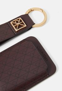Brązowy skórzany portfel na karty marki Calvin Klein z geometrycznym wzorem i dołączonym złotym brelokiem z kwadratowym emblematem.