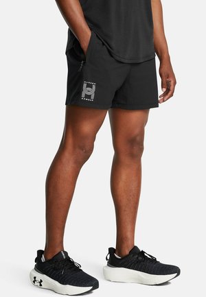 Mann trägt schwarze Under Armour Sportshorts und schwarze Laufschuhe mit weißen Sohlen, steht mit einer Hand in der Tasche.