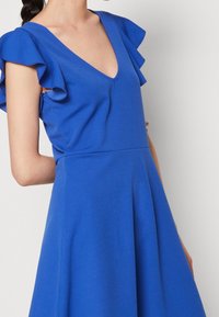 Kirkas sininen mekko, jossa on V-mallinen kaula-aukko ja röyhelöhihoja. Kangas näyttää sileältä, ja mekossa on leveä helma, joka ulottuu vyötäröltä.