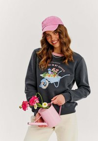 Sivá mikina s grafickým dizajnom kvetinového vozíka, skombinovaná s ružovou čiapkou. Drží ružový zálievkový kanister s kvetmi a stojí na bielom pozadí.