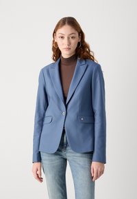 Světle modrý blazer z hladké tkaniny, zapínání na jeden knoflík, dvě přední kapsy a decentní logo na klopě, v kombinaci s hnědým topem.