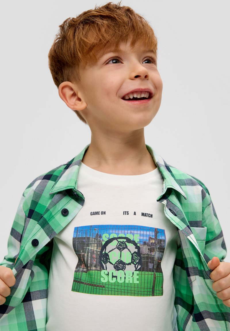 Zelena i plava košulja na kari preko bijelog T-shirta s grafikom nogometne lopte i podebljanim tekstom. Tekstura je glatka, ležeran kroj.