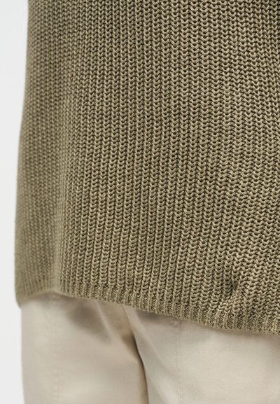 Κοντινό πλάνο της εσοχής ελιάς πράσινου πλεκτού πουλόβερ πάνω σε μπεζ παντελόνια, που δείχνει υφή πλεξίματος και λεπτομέρειες υφάσματος.