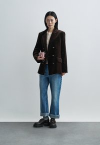 Pruun sametkostüüm, topeltreageeritud kuldsete nööpidega, kombineeritud heleroheliste mansetiga teksade ja mustade paksude kingadega, hoides väikest roosat kotti.