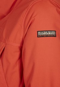 Oranssi synteettinen takkimateriaali, joka sisältää tikatun sauman ja mustan logo-eliippun, jossa on valkoiset tekstit "NAPAPIJRI geographic."