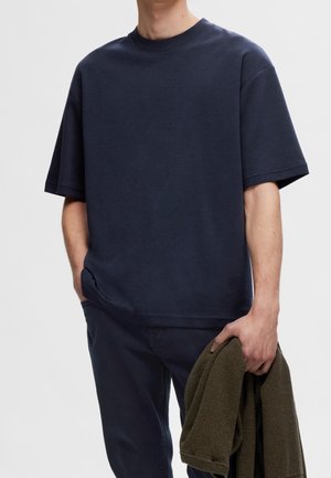Personne portant une chemise à manches courtes bleu marine et un pantalon, une main dans la poche et tenant un pull en tricot vert olive.