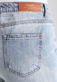 Světle modré džíny z denimu s odřeným vzhledem. Mají hnědou koženou visačku na zadním pásu a zadní kapsu s detaily prošívání.