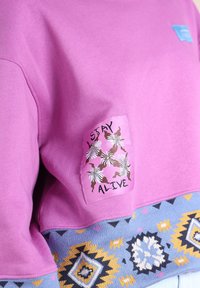 Sweatshirt violet avec un écusson brodé portant l'inscription "RESTEZ EN VIE", mettant en avant des motifs floraux. Le bas de la pièce présente un détail en tricot à motifs bleus.