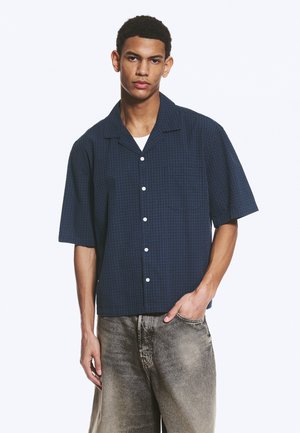 Jeune homme portant une chemise bleu marine à manches courtes boutonnée sur un sous-vêtement blanc avec un jean gris délavé, debout avec une main dans la poche.