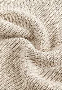 Beige gestrickter Stoff mit einem strukturierten Fischgrätmuster. Weiche, dicke Garne sorgen für einen gemütlichen und flexiblen Fall. Geeignet für Textilien und Polsterung.
