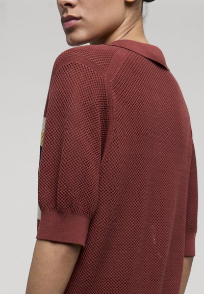 Femme portant un haut en maille texturée couleur rouille, avec des manches aux coudes et un col, vue de dos et de profil.