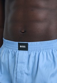 Svetlo modre bokserice iz mehkega materiala z elastičnim pasom. Črna nalepka z logotipom "BOSS" prišita na hrbtni strani.
