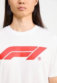 Baltas kokvilnas T-krekls ar lielu sarkanu Formula 1 logotipu, ar gludu audumu un noapaļotu izgriezumu.