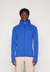 Chaqueta azul con cremallera y capucha, hecha de una tela suave y elástica. Cuenta con agujeros para los pulgares, bolsillos laterales y un diseño ajustado.