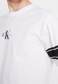 Valkoinen puuvillapaita pyöreällä kauluksella ja lyhyillä hihoilla, jossa on pieni musta Calvin Klein -logo ja mustat vaakasuorat raidat hihoissa.