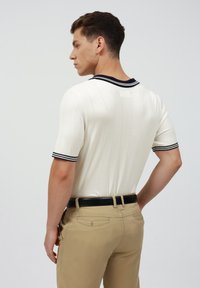 Polo de manga corta, de canalé, en blanco con cuello a rayas en azul marino. El modelo lleva pantalones caqui con un cinturón negro, visto desde atrás.
