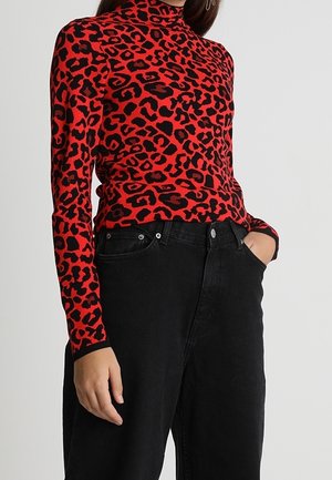 Femme portant un t-shirt col roulé rouge à manches longues avec imprimé léopard noir et un jean noir taille haute, une main dans la poche.