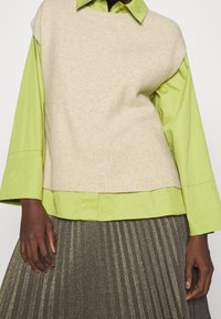 Dunkelhäutige Person, die eine hellbeige Strickweste über einem limettengrünen Shirt mit weiten Ärmeln und einem plissierten dunkelmetallischen Rock trägt.