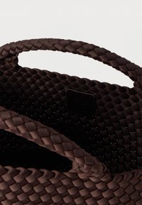 Καφέ υφαντή τσάντα χειρός με υφή μοτίβου, με δύο πλεκτές λαβές και μικρή ανάγλυφη ετικέτα με λογότυπο στο εσωτερικό.