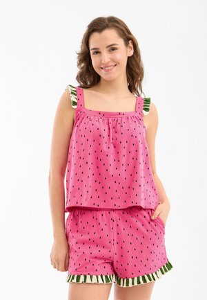 Mujer sonriendo, lleva un conjunto de top sin mangas y shorts rosa con puntos negros parecidos a semillas y ribete fruncido verde y blanco.