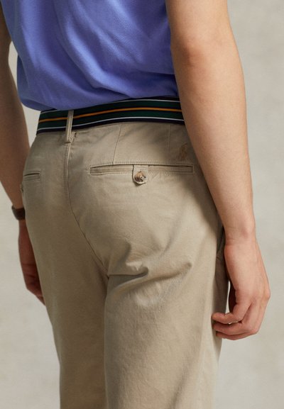 Persoană purtând pantaloni bej cu buzunar spate nasturat și curea în dungi, asortați cu o cămașă violetă băgată în pantaloni.