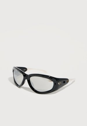 Occhiali da sole a occhi di gatto con montatura spessa nera, lenti trasparenti e stanghette bianche, posati su una superficie bianca.