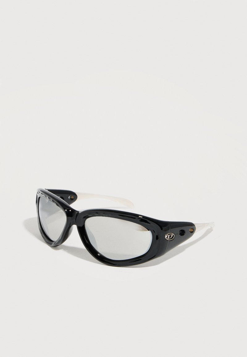 Gafas de sol estilo ojo de gato con montura negra gruesa, lentes transparentes y varillas blancas, colocadas sobre una superficie blanca.