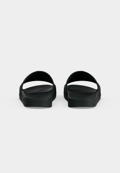 Ζευγάρι μαύρα σανδάλια τύπου slip-on, εμφανιζόμενα από πίσω με φαρδιές λουρίδες και επίπεδες μαξιλαρωτές σόλες σε απλό φόντο.