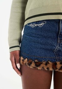 Denim mini skirt with leopard print faux fur trim, featuring embroidered branding. Green sweater with ribbed cuffs complements the look.